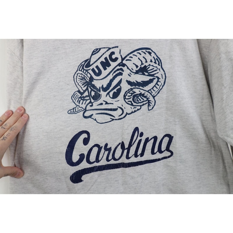 Op de afbeelding: Een lichtgrijs sweatshirt met een marineblauwe afbeelding van een rammenkop met de letters "UNC" op een hoed. Onder de afbeelding staat het woord "Carolina" in marineblauwe letters.