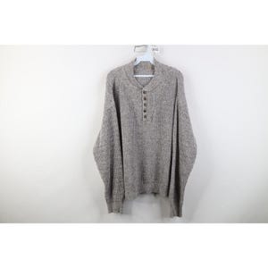 May include: A gray waffle-knit sweater with a henley neckline and four brown buttons. The long-sleeved sweater is hanging on a white hanger against a white background. The sweater has a relaxed fit.