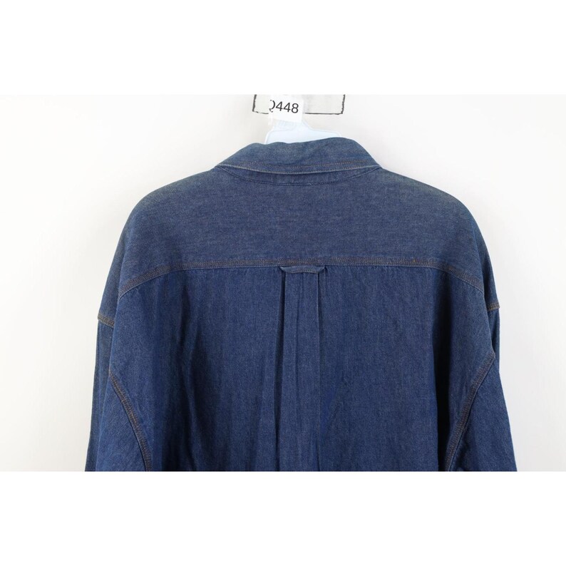 May include: A close-up of the back of a blue denim shirt. The shirt has a collar, a yoke, and a vertical pleat down the center. The stitching is visible in a contrasting color. The shirt is hanging on a white background.
