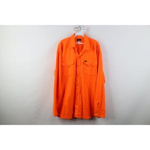 May include: Bright orange long-sleeve button-down shirt with two chest pockets and a small black label. The shirt is hanging on a white hanger against a white background. The shirt has a classic collar and buttoned cuffs.