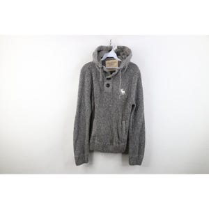 May include: A gray knit hooded sweater with a button-up neckline and a kangaroo pocket. The sweater features a small white logo on the chest and is hanging on a white hanger. The label reads "Abercrombie & Fitch".