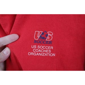 May include: Close-up of a red garment featuring the US Soccer Coaches Organization logo. The logo includes the letters "US" in a white outline, with a soccer ball graphic, and the word "SOCCER" below. The text "US SOCCER COACHES ORGANIZATION" is also visible.