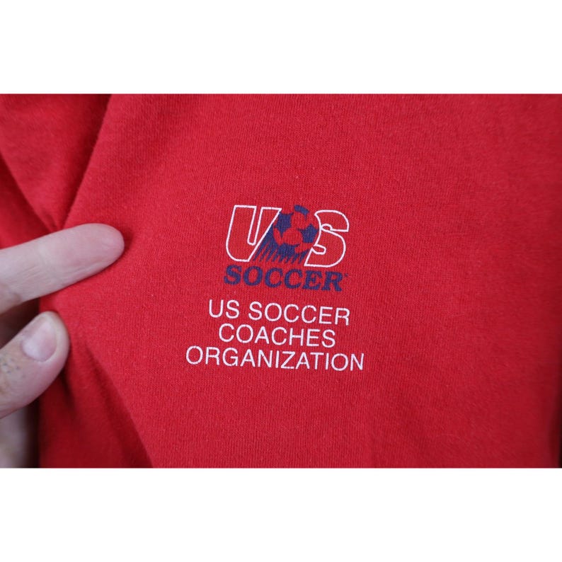 May include: Close-up of a red garment featuring the US Soccer Coaches Organization logo. The logo includes the letters "US" in a white outline, with a soccer ball graphic, and the word "SOCCER" below. The text "US SOCCER COACHES ORGANIZATION" is also visible.