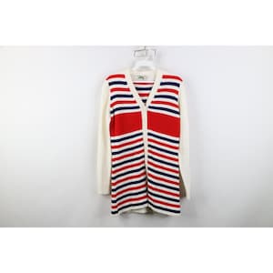 May include: A long, cream-colored cardigan with a V-neck and button closure. The front features a bold design of red and navy blue horizontal stripes. The sleeves are solid cream, and the sweater hangs on a white hanger.
