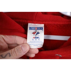 Peut inclure: Gros plan d'un sweat &agrave; capuche rouge avec une &eacute;tiquette de la marque Jerzees. L'&eacute;tiquette affiche le logo de la marque, des informations de taille (XL, XG, TG) et la composition du tissu (50% coton, 50% polyester). L'&eacute;tiquette indique &eacute;galement que l'article a &eacute;t&eacute; fabriqu&eacute; au Honduras.