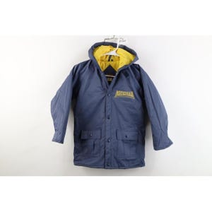 Puede incluir: Una chaqueta con capucha azul marino que presenta un forro interior amarillo brillante. En la parte delantera se muestra la palabra MICHIGAN bordada en hilo amarillo. Incluye cierre de botones a presión, mangas largas y dos bolsillos frontales con solapa.