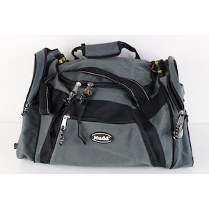 May include: A grey and black duffel bag featuring multiple zippered compartments, black webbing handles, and a padded top handle. The front displays a black oval patch with the white text Mudd. The bag includes silver-toned metal zipper pulls.