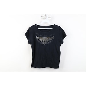 May include: Black Harley-Davidson t-shirt featuring a silver and gold winged graphic and the brand name. The short-sleeved top is displayed on a white hanger against a white backdrop. The shirt has a crew neck.