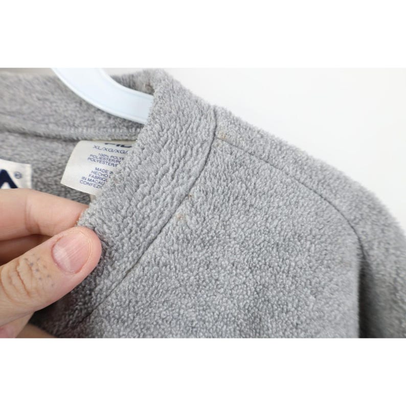 May include: Close-up of a gray fleece jacket. The jacket has a soft, textured surface and a high collar. A tag with text is visible. The jacket is held by a hand.