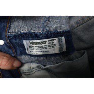 May include: Close-up of a Wrangler jeans label. The label is white with black text, indicating the size as 33 x 34 inches. The label also includes washing instructions and the fabric composition. The jeans are made in Mexico of US fabric.
