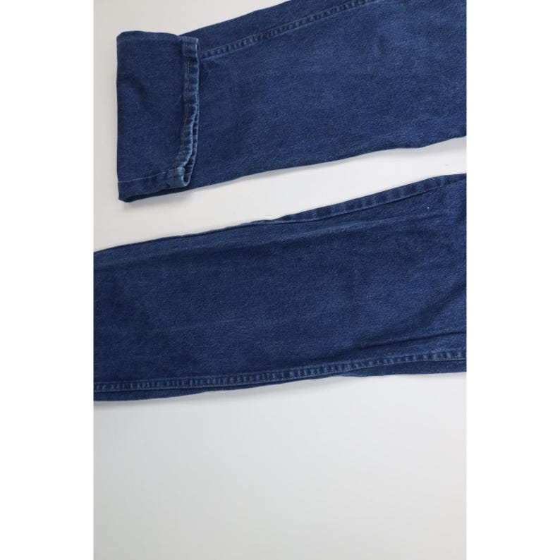 May include: Close-up of a pair of dark blue denim jeans. The jeans are folded at the cuff, showing the detailed stitching. The fabric has a classic, slightly faded appearance.
