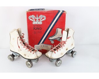 May include: A pair of off-white roller skates with red laces and metal wheels, positioned in front of a red and white cardboard box labeled Street King Out Door Shoe Skates with a crown graphic and a diagonal blue stripe across the front.