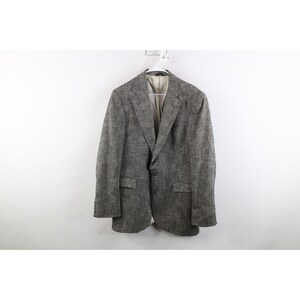 May include: A gray and black textured blazer featuring a notched lapel, two front buttons, two flap pockets, and long sleeves with buttoned cuffs. The garment has a light beige interior lining and is displayed on a white plastic hanger.