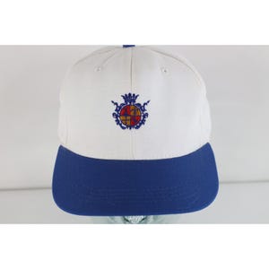 May include: A white baseball cap with a solid blue brim. The front panel features an embroidered crest in blue, red, and yellow, depicting a crown above a shield divided into four quadrants with a fleur-de-lis symbol in the upper right section.