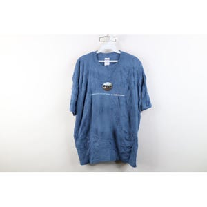 Op de afbeelding: Een blauw t-shirt met korte mouwen en een cirkelvormig Dell-logo op de borst. Onder het logo staat de tekst: GET MORE OUT OF YOUR ENTERPRISE. GET MORE OUT OF NOW. Het shirt heeft een ronde hals en hangt aan een witte plastic kledinghanger.
