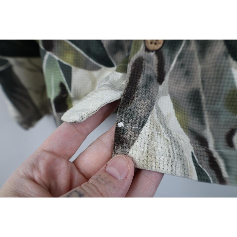 Op de afbeelding: Close-up van een overhemd met knopen en een patroon. Het overhemd heeft een mix van groene, bruine en beige kleuren in een textuurstof. Een houten knoop is zichtbaar bij de kraag.