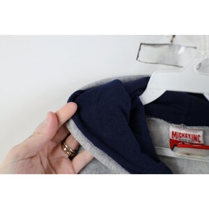 May include: A heather gray hooded sweatshirt featuring a dark navy blue interior hood lining. A white fabric tag with red text reading MICKEY INC SINCE 1928 is stitched into the inner neckline of the garment, which is displayed on a white hanger.
