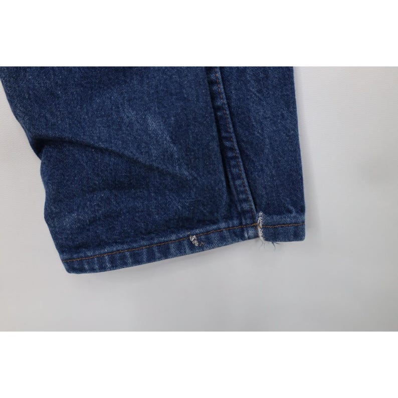 Puede incluir: Primer plano del dobladillo de unos vaqueros de mezclilla azules. La mezclilla es de un lavado medio con costuras visibles y un ligero deshilachado en el dobladillo. La tela est&aacute; doblada, mostrando la textura y las variaciones de color.