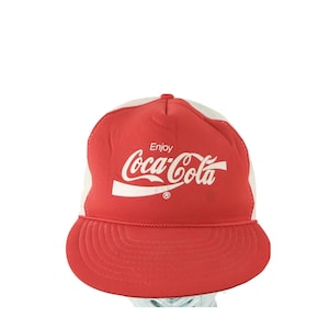 May include: Red and white Coca-Cola trucker hat. The hat features a red front panel with the Coca-Cola logo in white, a white mesh back, and a red brim. The text "Enjoy Coca-Cola" is printed on the front.