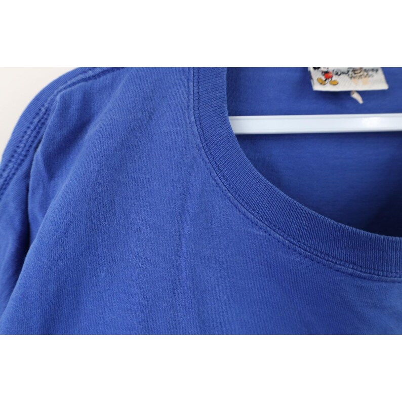 May include: Close-up of a blue t-shirt with a crew neck. The shirt has a tag that reads "Walt Disney World". The shirt is hanging on a white hanger.