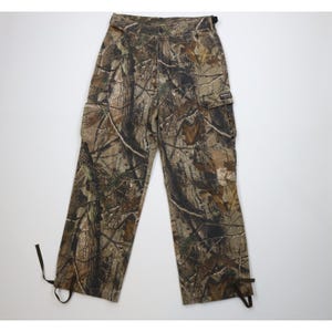 May include: Camouflage cargo pants in shades of brown, green, and tan. The pants feature multiple pockets, a button and zipper closure, and adjustable ties at the ankles. The fabric has a realistic forest pattern.