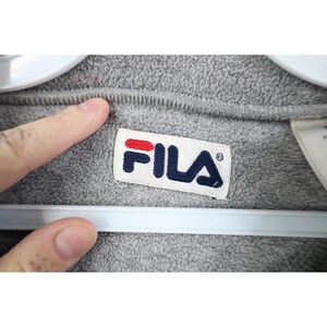 May include: Close-up of a gray fleece garment with a white label featuring the FILA logo in red and navy blue. The garment is hanging on a white hanger.