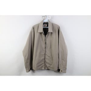 Puede incluir: Chaqueta beige con cremallera completa, cuello y puños abotonados. La chaqueta está colgada de una percha blanca sobre un fondo blanco. La chaqueta tiene un pequeño logotipo en el pecho.