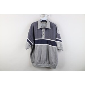 Könnte beinhalten: Ein kurzärmeliges Poloshirt mit einem Color-Block-Design, bestehend aus einem grauen, strukturierten Oberteil, einem dunkelblauen horizontalen Bruststreifen mit weißen Linien und einem einfarbigen grauen Unterteil. Es verfügt über einen Kragen, eine Knopfleiste mit drei Knöpfen und einen Saum.