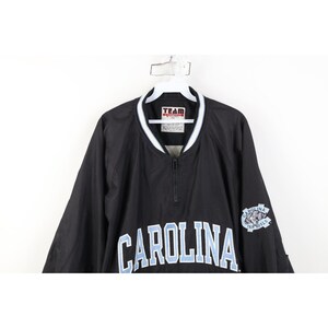 May include: Black pullover jacket with a white and light blue collar and the word "CAROLINA" across the chest in light blue lettering. The jacket has a small logo on the right sleeve.