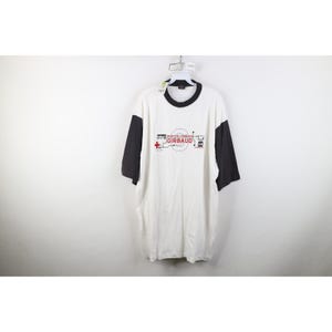May include: White t-shirt with black sleeves and collar. The shirt has a graphic design with the text "MARITIME FRANCOIS GIRBAUD" in black and red. The shirt is hanging against a white background.