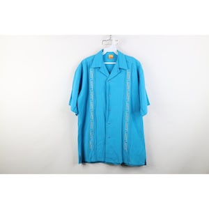 May include: A turquoise short-sleeved shirt with a camp collar. The shirt has vertical embroidered designs on the front. It is hanging on a white hanger against a white background. The shirt has a relaxed fit and is buttoned up.