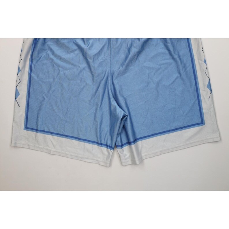May include: Light blue athletic shorts featuring a solid light blue center panel framed by a darker blue rectangular border. The side panels are white with a black diamond argyle pattern, and the bottom hem is finished with a wide white band.