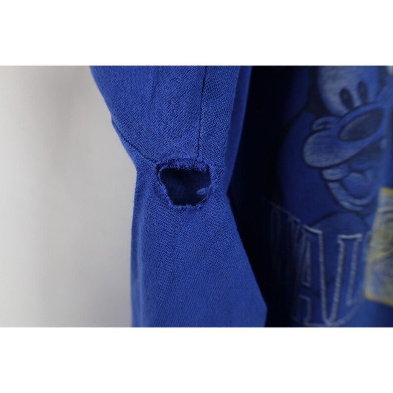 May include: Close-up of a royal blue t-shirt with a distressed hole near the shoulder seam. The right side of the shirt features a cartoon character graphic. The shirt appears to be vintage.