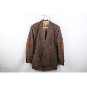 May include: Brown tweed blazer with suede elbow patches. The jacket has two front pockets and a two-button closure. The blazer is hanging on a white hanger against a white background.