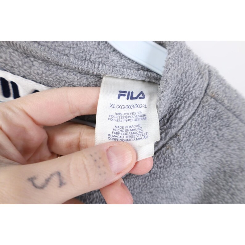 May include: Close-up of a gray FILA fleece jacket. The tag reads "XL/XG/XG/XG/XL" and lists the material as 100% polyester. The tag also indicates the jacket was made in Macau.