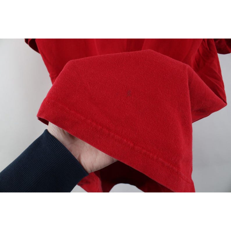 May include: Close-up of a bright red t-shirt. The fabric is folded, revealing the hem and a small dark spot. The sleeve is visible in the background. The shirt is held by a person wearing a navy blue long-sleeve shirt.