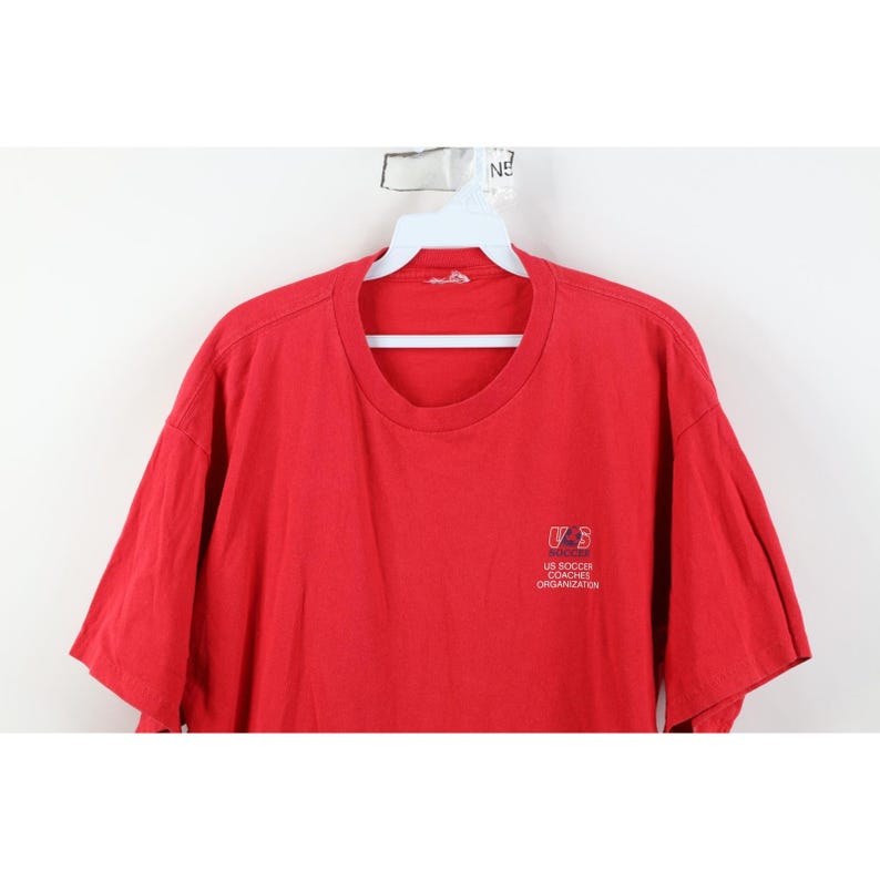 May include: A red t-shirt with a crew neck and short sleeves. The shirt features a small logo on the upper left chest that reads "US Soccer Coaches Organization". The shirt is made of a soft, likely cotton material.