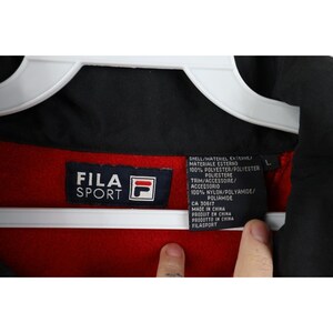 May include: Close-up of a black, red, and white Fila Sport jacket tag. The tag includes the Fila Sport logo, material information, and the text "MADE IN CHINA". The jacket is hanging on a white hanger.