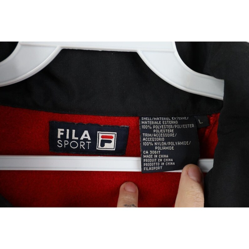 May include: Close-up of a black, red, and white Fila Sport jacket tag. The tag includes the Fila Sport logo, material information, and the text "MADE IN CHINA". The jacket is hanging on a white hanger.