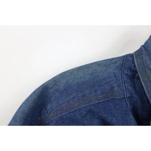 May include: Close-up of a blue denim jacket shoulder and collar. The jacket has visible stitching in a contrasting brown thread. The denim has a faded, worn appearance, suggesting it has been washed.