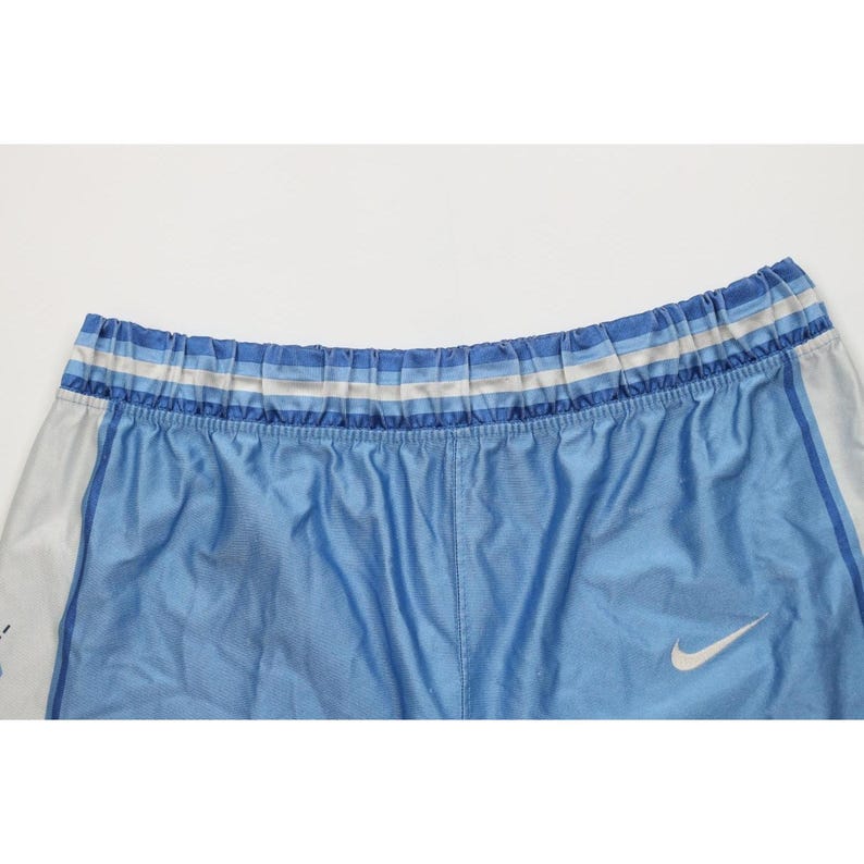 May include: Light blue athletic shorts featuring a white Nike swoosh logo on the lower right side. The waistband has a horizontal striped pattern in shades of blue and white. The sides of the shorts have white panels with blue trim.