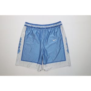 May include: Light blue and white athletic shorts featuring an elastic waistband with horizontal stripes. The front panels are light blue with a white Nike swoosh logo, while the side panels display a dark blue argyle pattern on a white background.