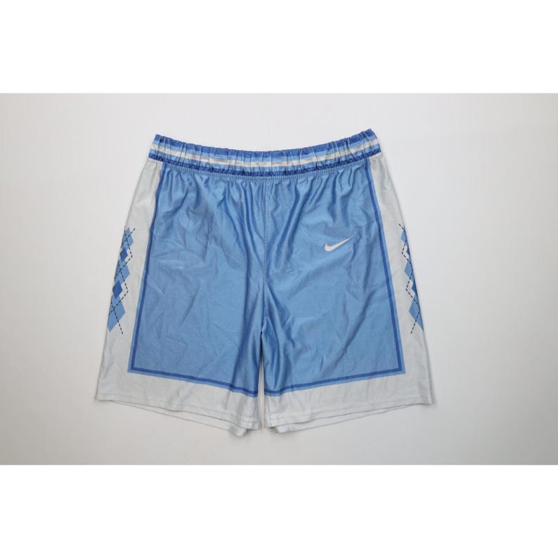 May include: Light blue and white athletic shorts featuring an elastic waistband with horizontal stripes. The front panels are light blue with a white Nike swoosh logo, while the side panels display a dark blue argyle pattern on a white background.