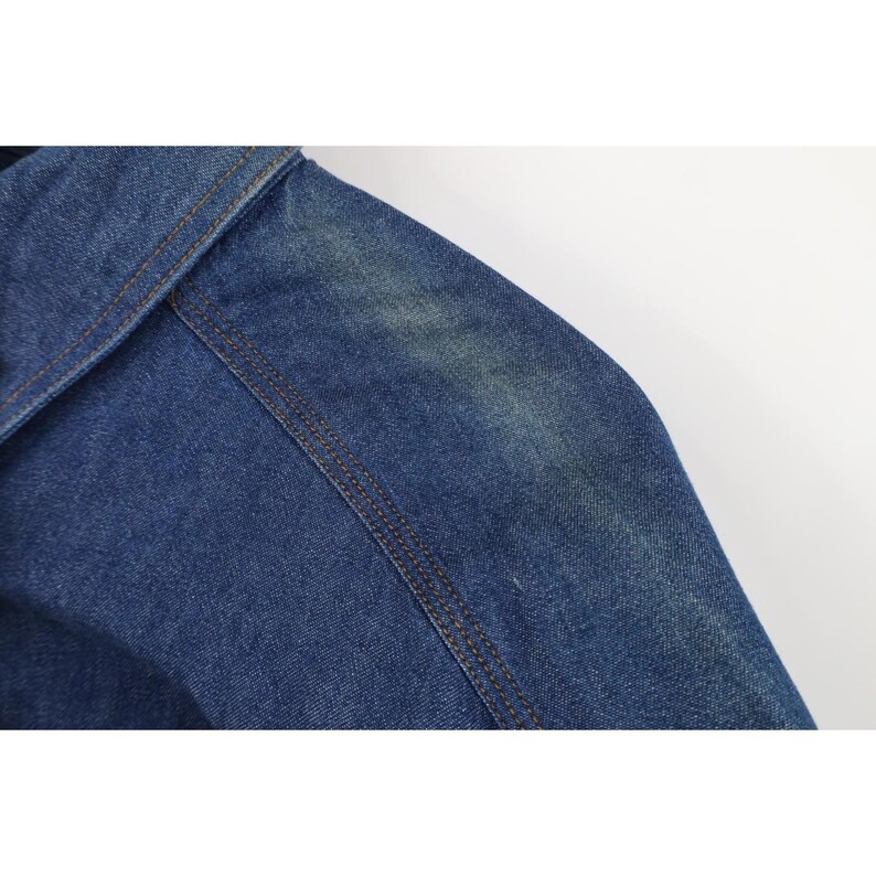 May include: Close-up of a blue denim jacket shoulder and sleeve. The jacket features visible brown stitching along the seams and a slightly faded appearance, suggesting wear. The background is plain white.