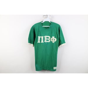 Könnte beinhalten: Ein grünes Kurzarm-T-Shirt mit weißen griechischen Buchstaben auf der Brust. Das Shirt hat einen Rundhalsausschnitt und weiße Bündchen an den Ärmeln. Ein kleines weißes rechteckiges Etikett ist am unteren linken Saum des Kleidungsstücks sichtbar.