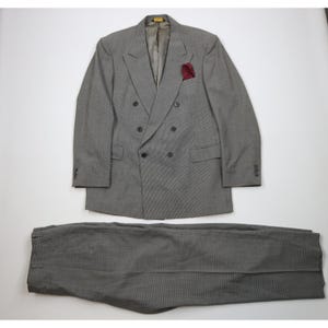 May include: A gray double-breasted suit with a matching pair of trousers. The suit features a red pocket square and black buttons. The suit is made of a textured fabric and is laid out on a white surface.
