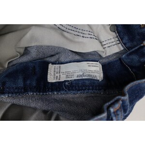 Puede incluir: Primer plano de unos vaqueros Wrangler de mezclilla azul. Los vaqueros tienen una etiqueta blanca con el logotipo de Wrangler y la informaci&oacute;n de la talla. Los vaqueros son 100% algod&oacute;n y miden 29x34 pulgadas (73,66 x 86,36 cm).