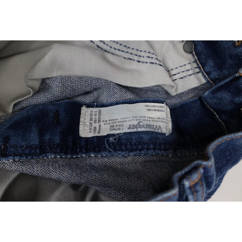 Puede incluir: Primer plano de unos vaqueros Wrangler de mezclilla azul. Los vaqueros tienen una etiqueta blanca con el logotipo de Wrangler y la informaci&oacute;n de la talla. Los vaqueros son 100% algod&oacute;n y miden 29x34 pulgadas (73,66 x 86,36 cm).