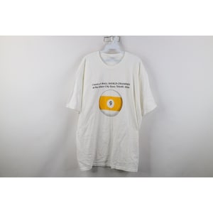 May include: White t-shirt with a graphic of a yellow and white 9-ball. The text above the graphic reads "I beat a 9 BALL WORLD CHAMPION at the Glass City Open, Toledo, Ohio."
