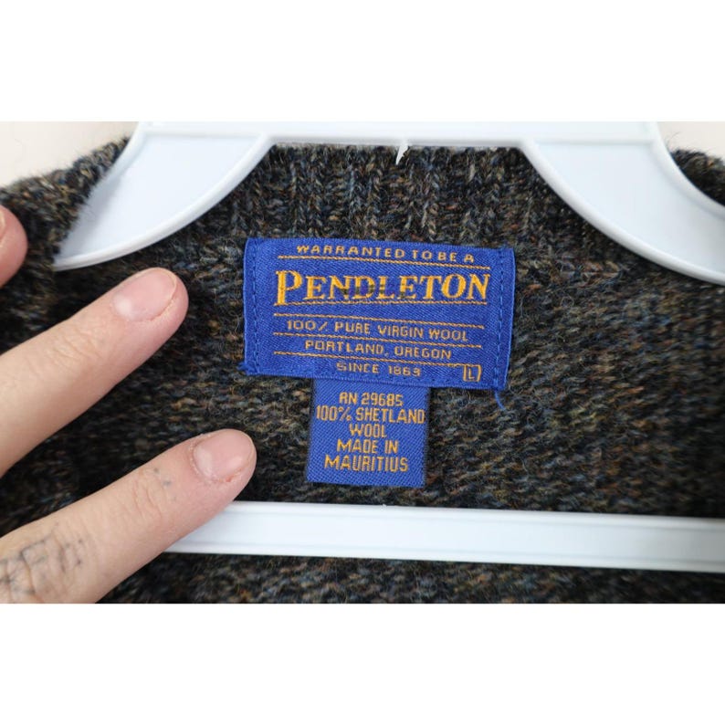 May include: Close-up of a Pendleton wool sweater with a blue label. The label reads "WARRANTED TO BE A PENDLETON" and "100% PURE VIRGIN WOOL". The sweater is a mix of brown, green, and black colors. The label also states "MADE IN MAURITIUS".